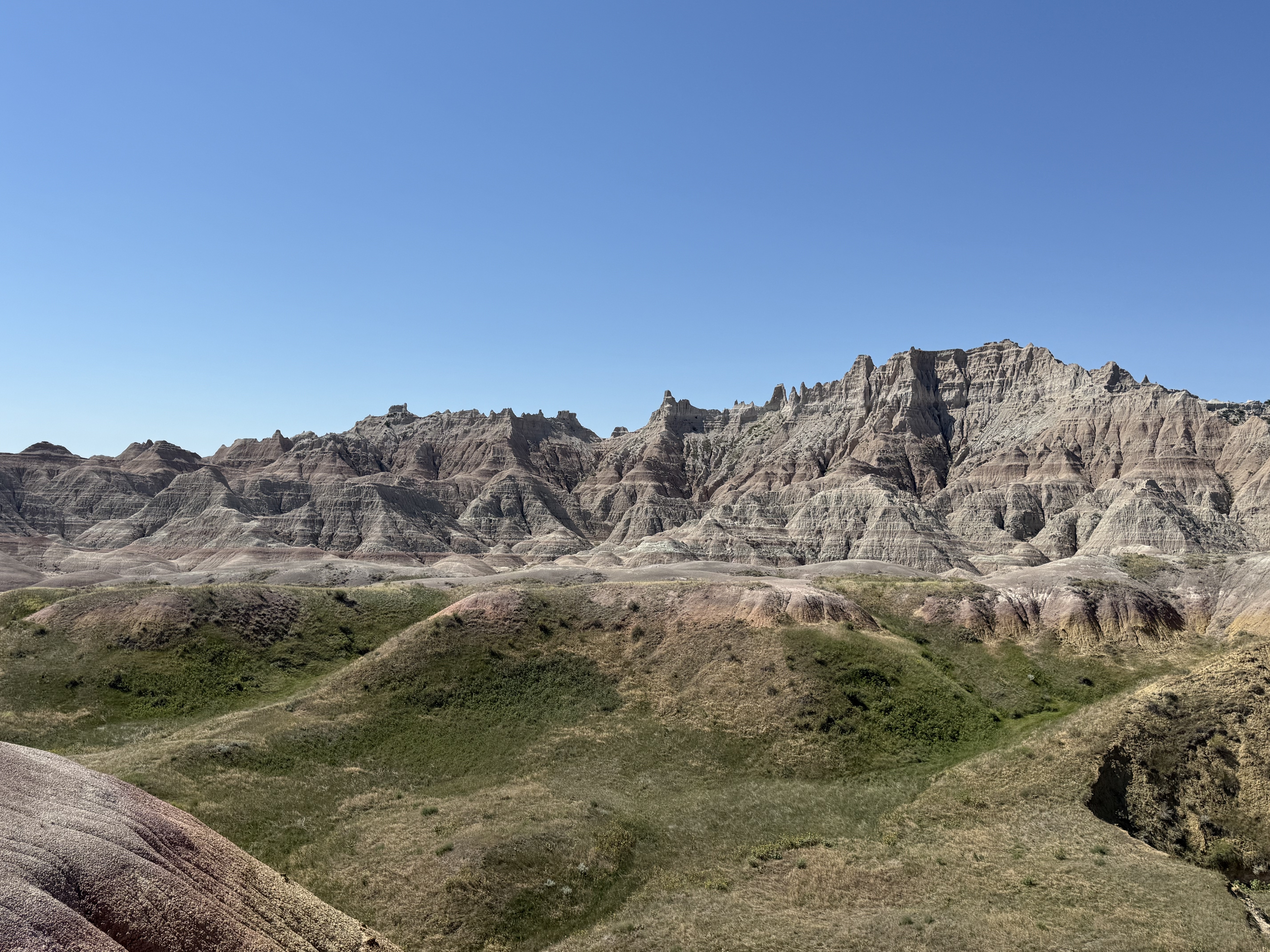 Rugged, layered badlands with green, rolling hills under a clear blue sky.