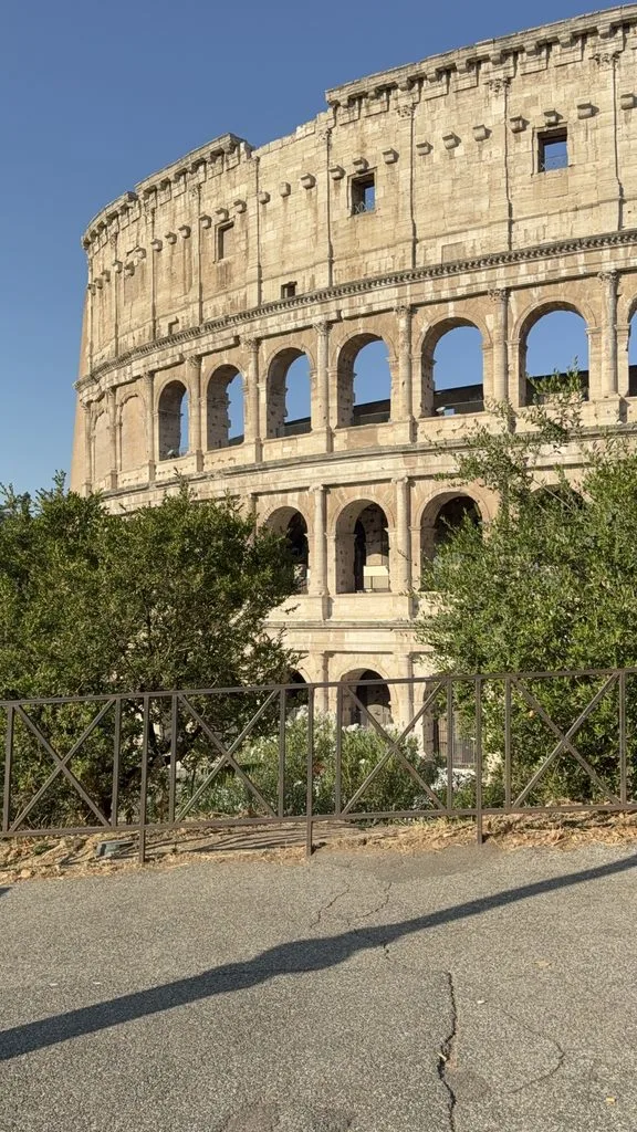 The Colosseum’s weathered stone arches and upper walls rise behind trees and a metal fence under a clear blue sky.